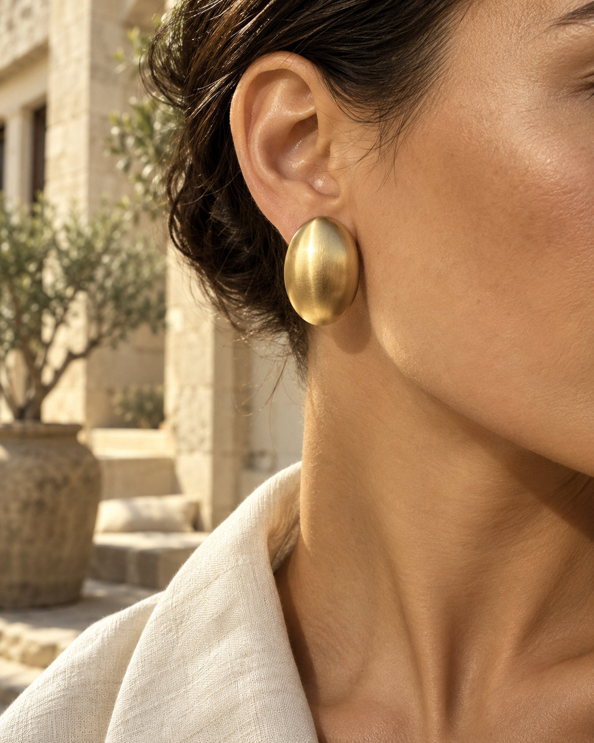 Femme portant une boucle d'oreille ovale dorée brossée, olivier et col en lin derrière, gros plan