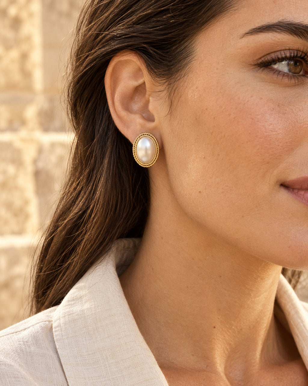 Woman wearing an oval pearl stud earring with gold rope border, close-up profile by a stone wall