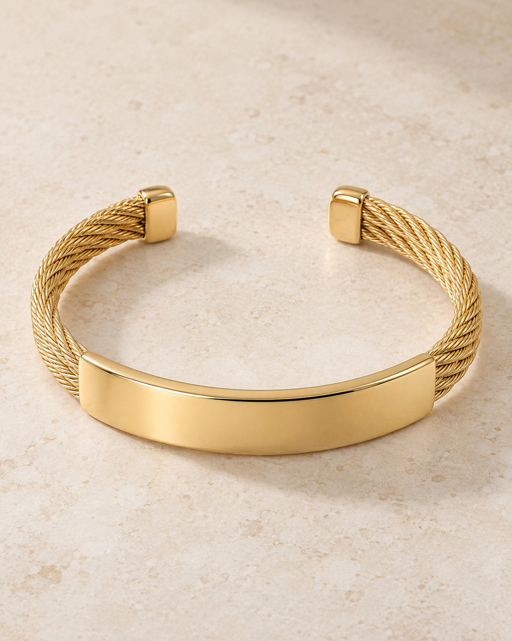 Gold-tone cable open cuff bracelet with smooth plate, shown on a travertine stone surface