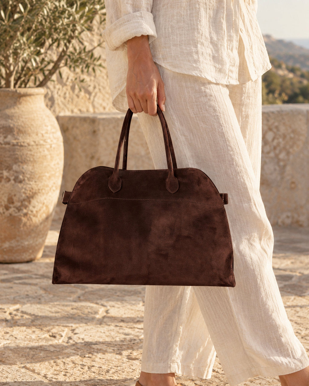 Dark brown suede-look arched handbag carried by hand with cream linen trousers on a stone terrace